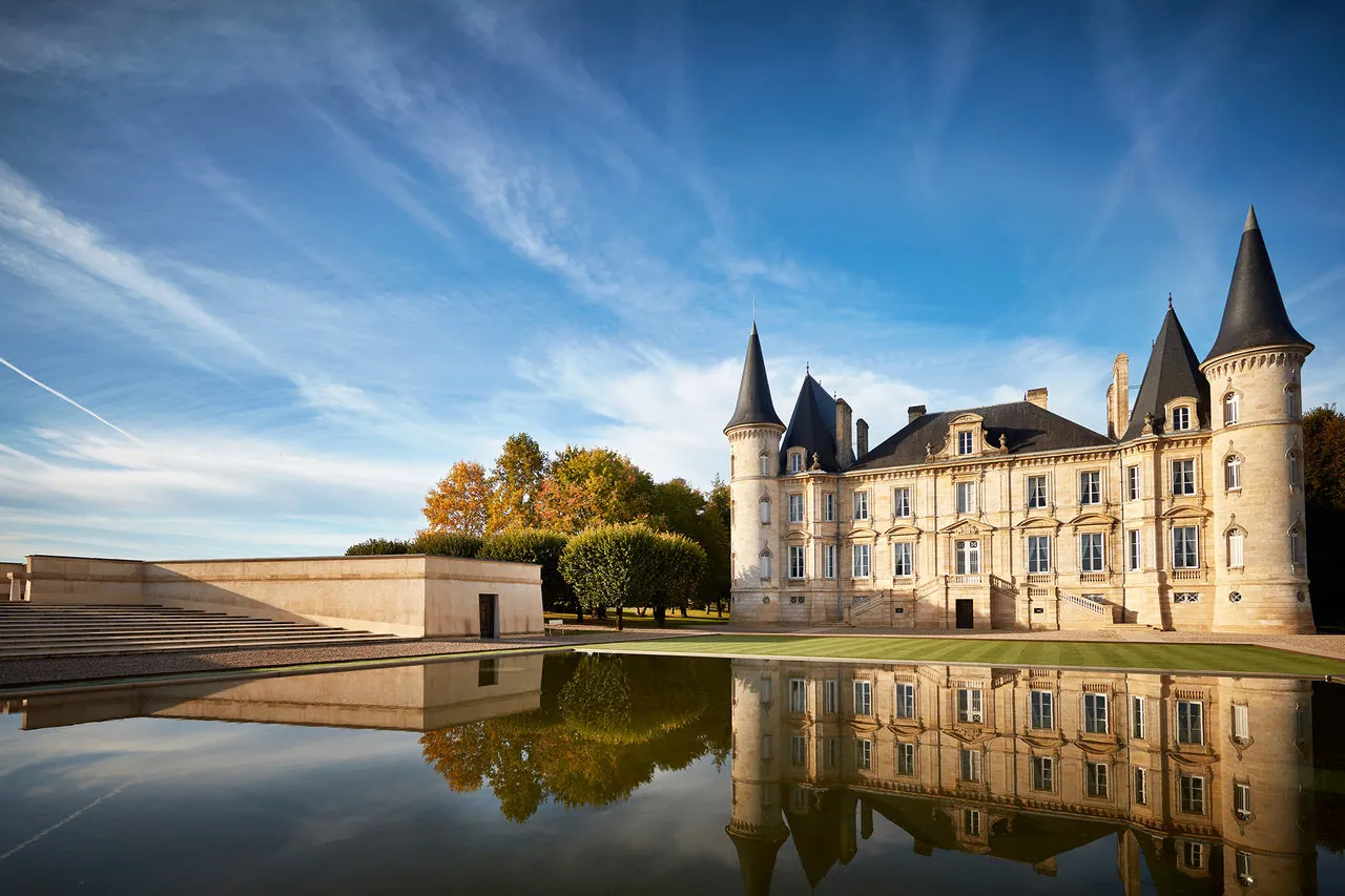 Château Pichon Baron in Bordeaux featuring distinctive conical towers and stone façades, reflected in still water, surrounded by manicured gardens and outbuildings