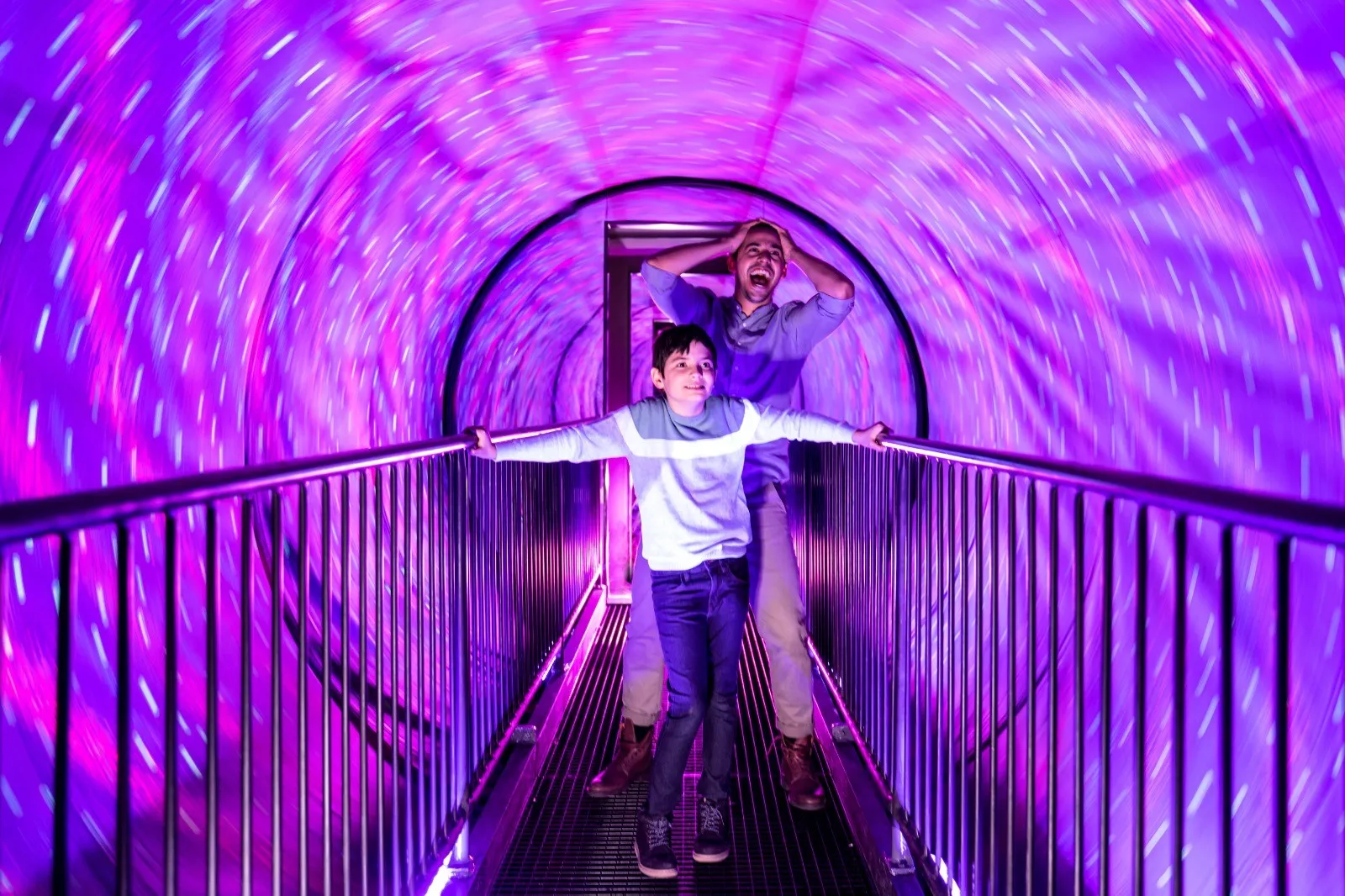 Two children in a neon pink and purple illuminated tunnel at the Museum of Illusions in Bordeaux, one with arms outstretched and the other raising their hand in joy.