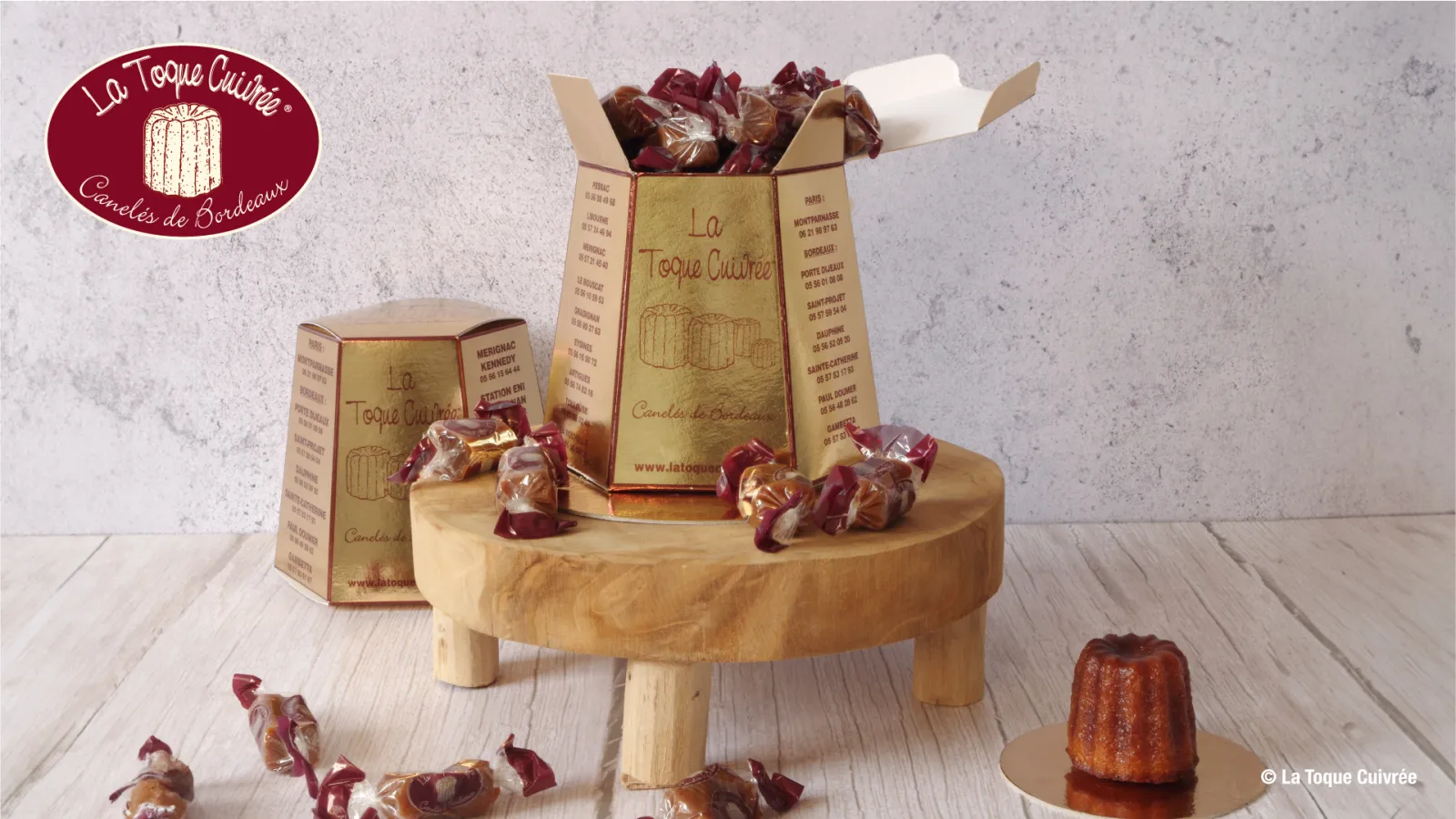Presentation of Canelés de Bordeaux by La Toque Cuivrée: golden cardboard boxes, canelés wrapped in burgundy and gold paper, and a single canelé on a round wooden display stand, with the brand logo in the top left.