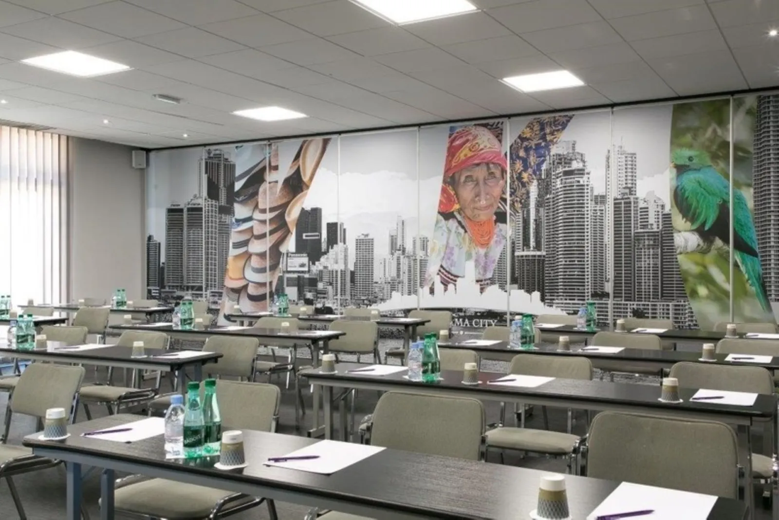 Modern conference room at Mercure Bordeaux Airport Hotel featuring black tables, beige chairs, and accent wall with urban cityscape collage including skyscrapers, portrait, and green parrot imagery.