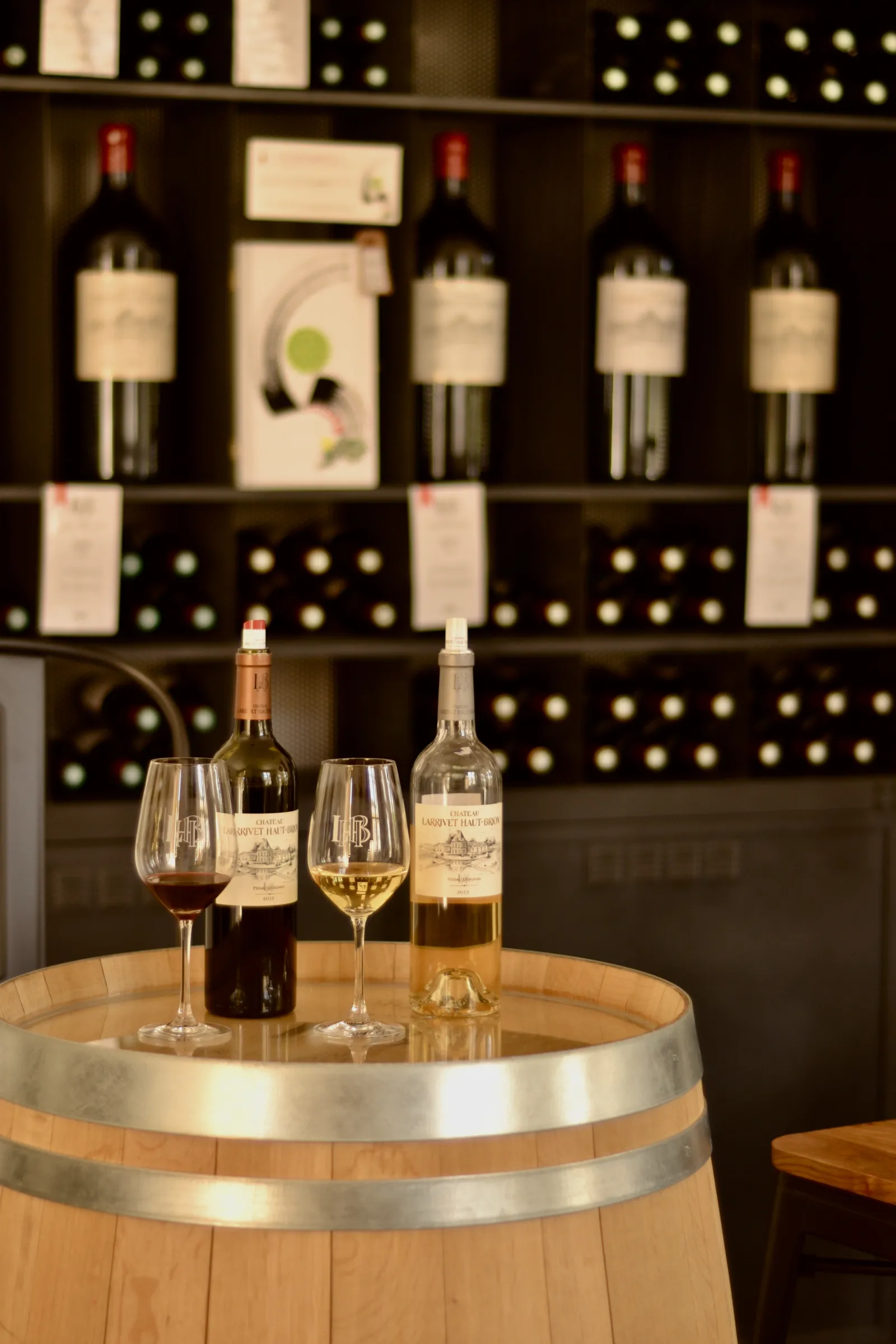 Wine tasting at Château Larrivet Haut-Brion in Bordeaux: red and white wine glasses on a barrel with bottles and wine cellar storage in the background.