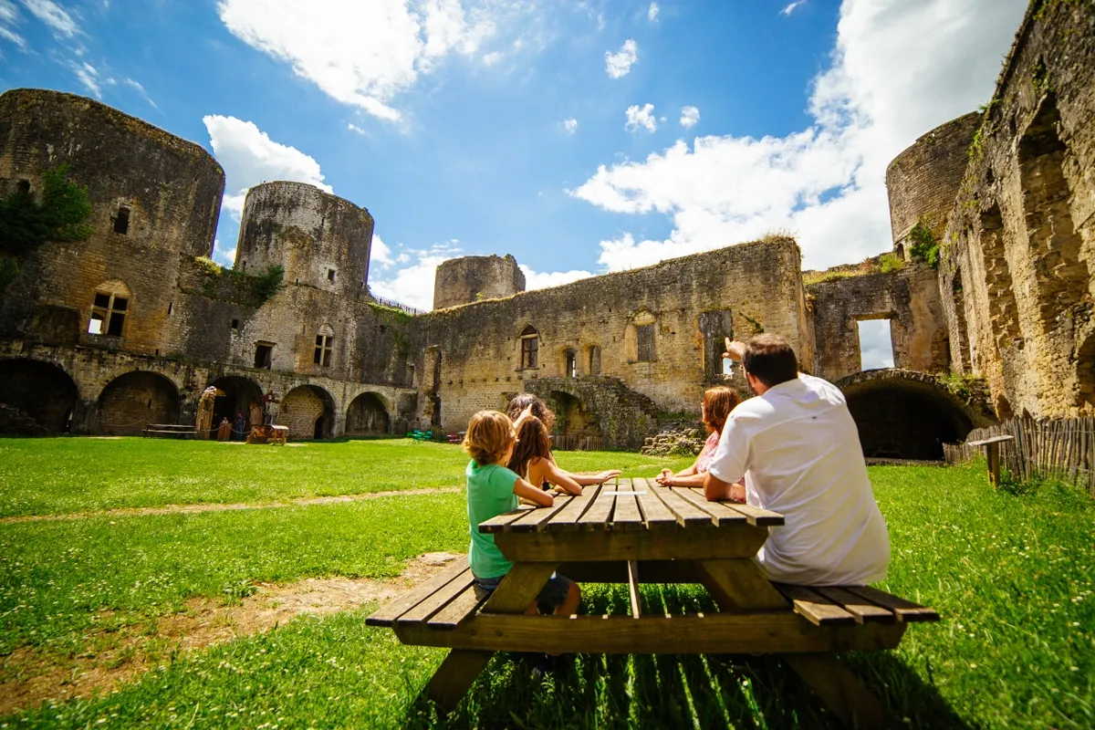 A family enjoys a picnic at a wooden table in the grassy courtyard of Villandraut Castle, surrounded by medieval stone walls and towers under a blue sky.