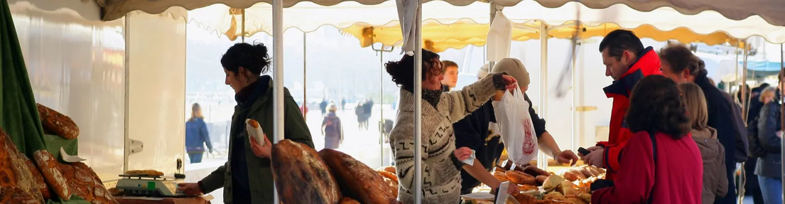Baker displaying artisanal bread under a market tent, serving customers selecting fresh bakery products.