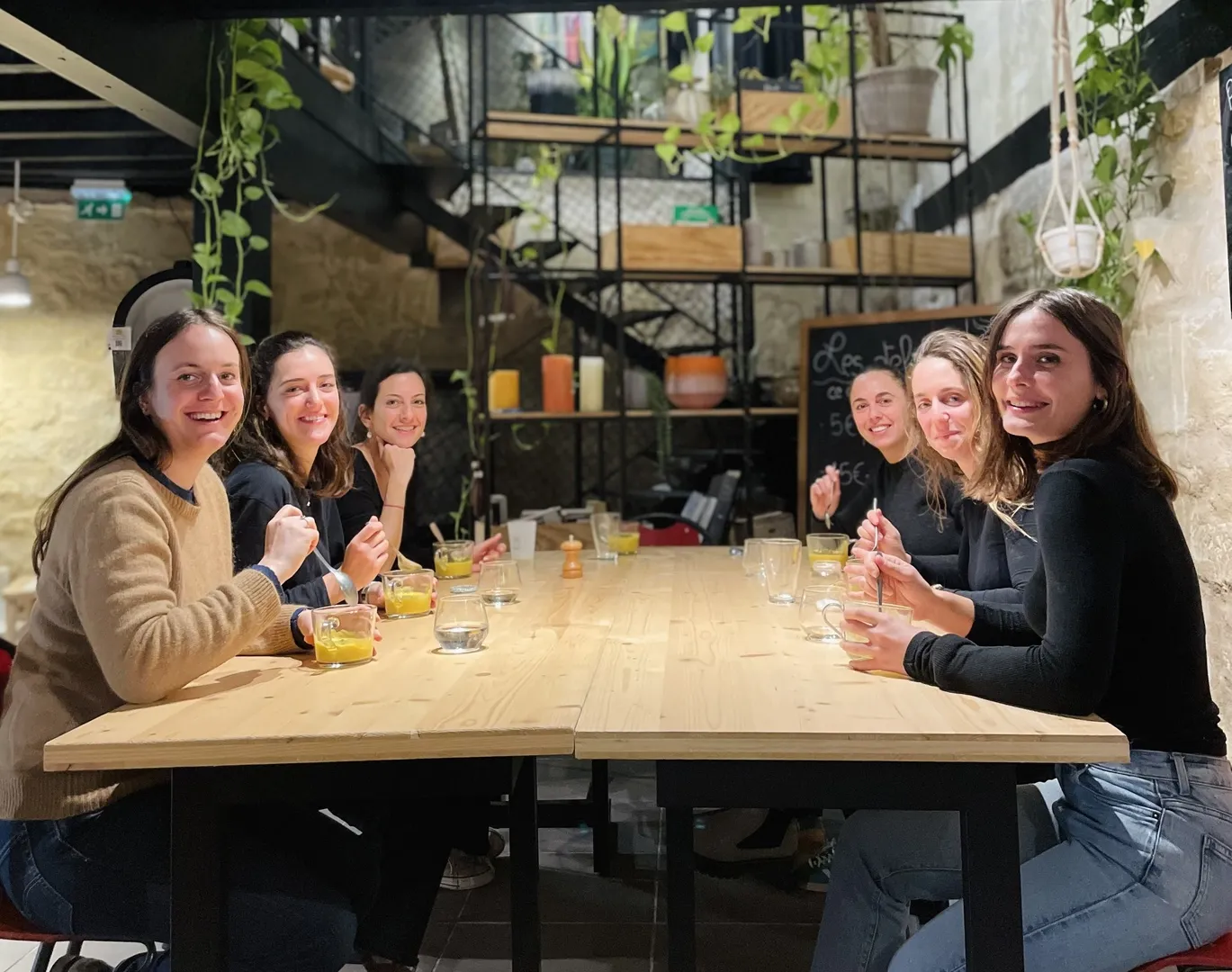 Seven young women sitting around a wooden table in a modern Bordeaux restaurant, smiling at the camera with glasses of water, in an industrial setting with hanging plants and metal shelves.