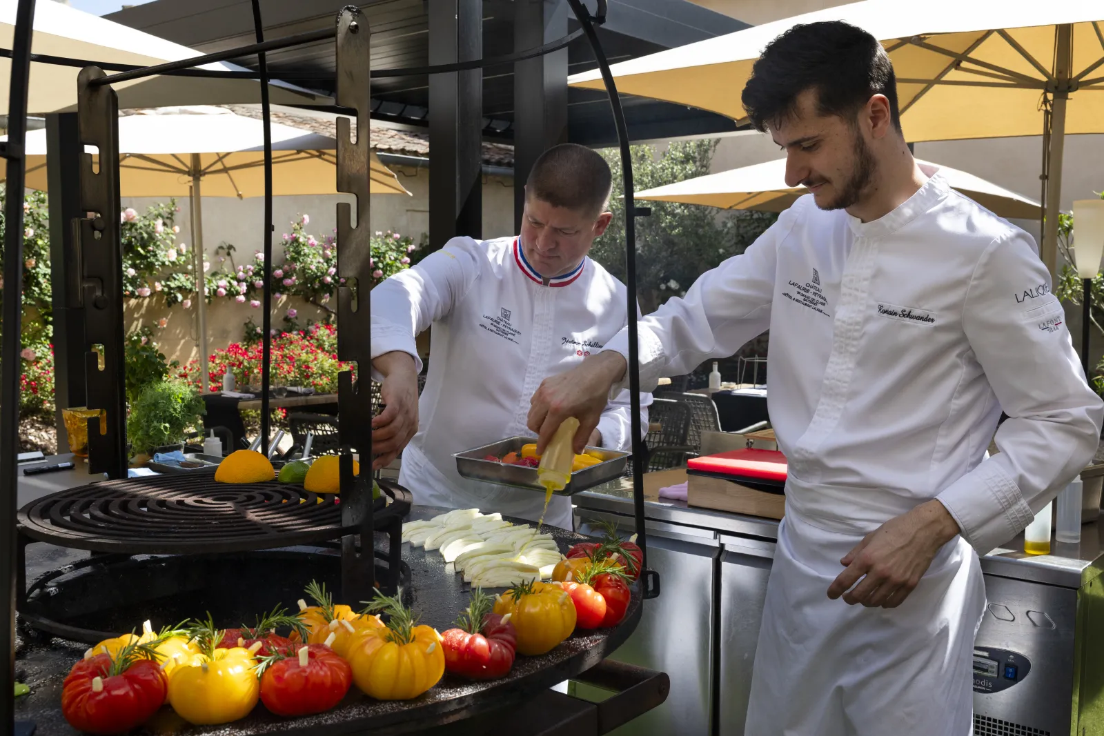 Two professional chefs in white chef's coats prepare fresh vegetables (tomatoes and zucchini) on a shaded terrace with yellow umbrellas at Bordeaux, La Terrasse de Lafaurie.