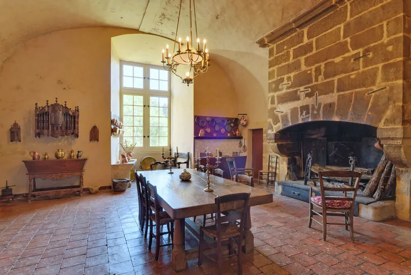 Dining room of the Royal Castle of Cazeneuve featuring a wooden table, rush-seat chairs, large brick fireplace, golden chandelier, and ochre-yellow walls.