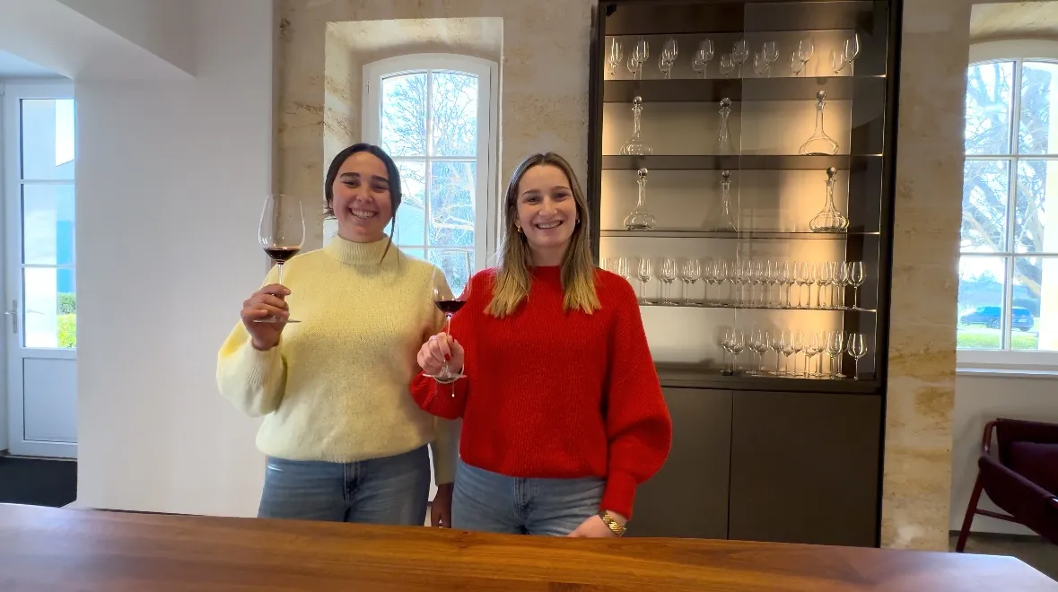 Two smiling young women in a Bordeaux wine tasting cellar, one wearing a yellow sweater and the other a red sweater, holding red wine glasses, with an illuminated glass display cabinet in the background and vineyard views through the windows.