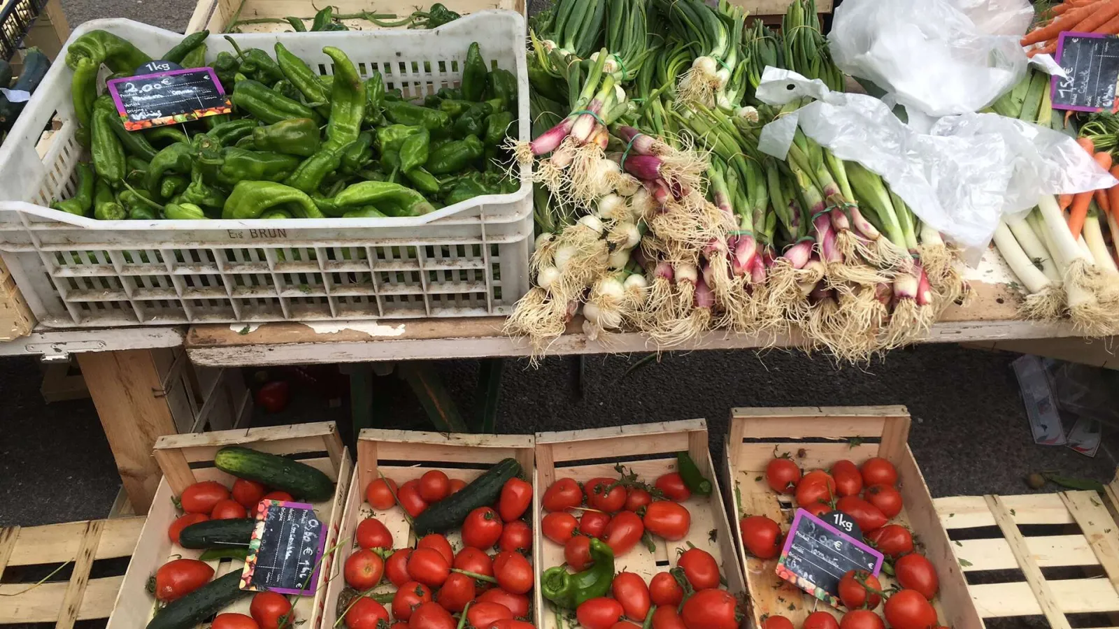 Market stall displaying fresh vegetables: green peppers in white crates, red onions with stems, tomatoes and cucumbers in wooden crates, at Eysines market in Bordeaux.