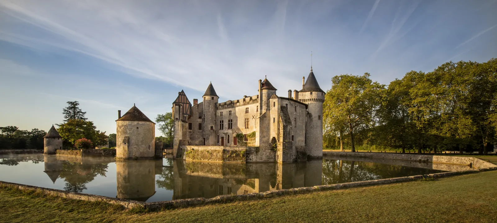 Château de La Brède near Bordeaux, medieval fortress with pointed towers surrounded by moats, reflected in still water, surrounded by trees and green fields under clear sky.