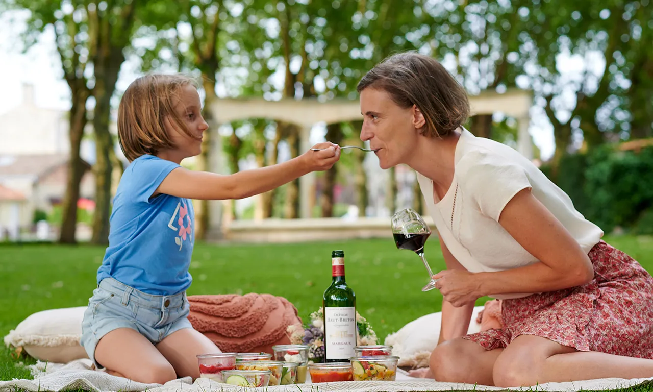 A woman and child enjoying a picnic in a park in Bordeaux, with the woman holding a glass of red wine while the child offers her a spoon, surrounded by wine bottle and preserved foods on a blanket.