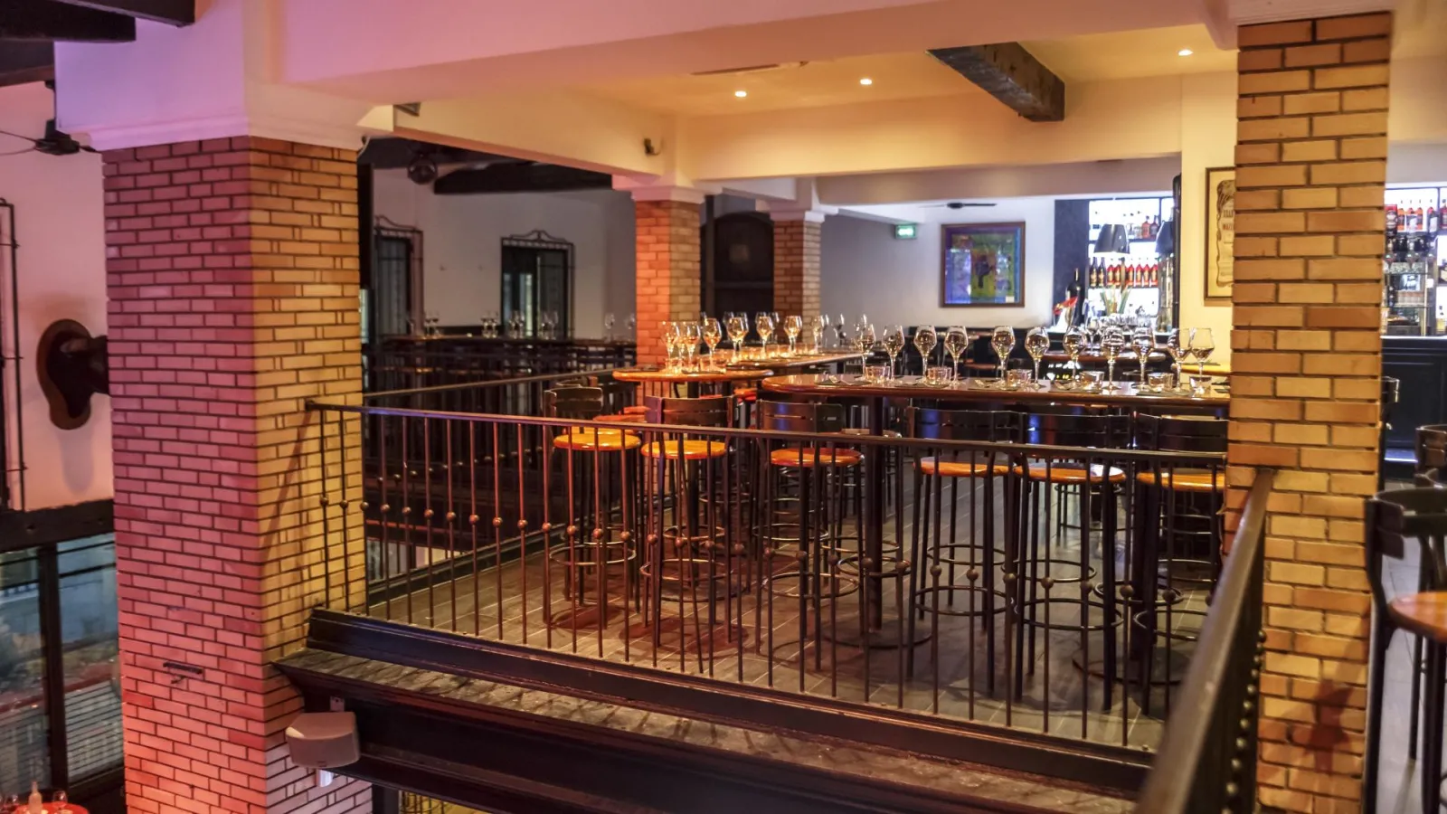 Interior view of Bodega Bodega bar in Bordeaux featuring wooden counter with wine glasses, red brick walls, warm pink and gold lighting, and black metal railings.