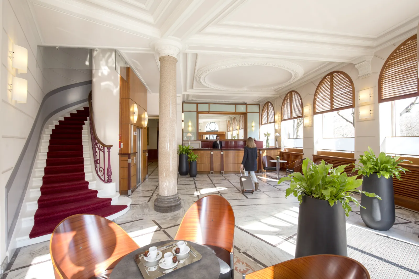 Elegant lobby of Hôtel de Normandie in Bordeaux featuring red marble staircase, wooden reception desk, white columns, green plants and guests