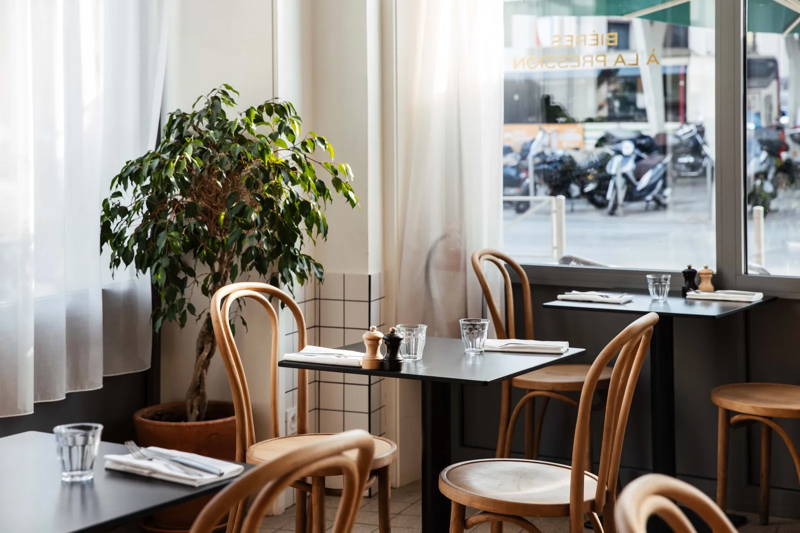 Elegant interior of Le Saint-Jean bistro in Bordeaux featuring black tables, bentwood chairs, potted plant, large windows overlooking the street, and minimalist decor.