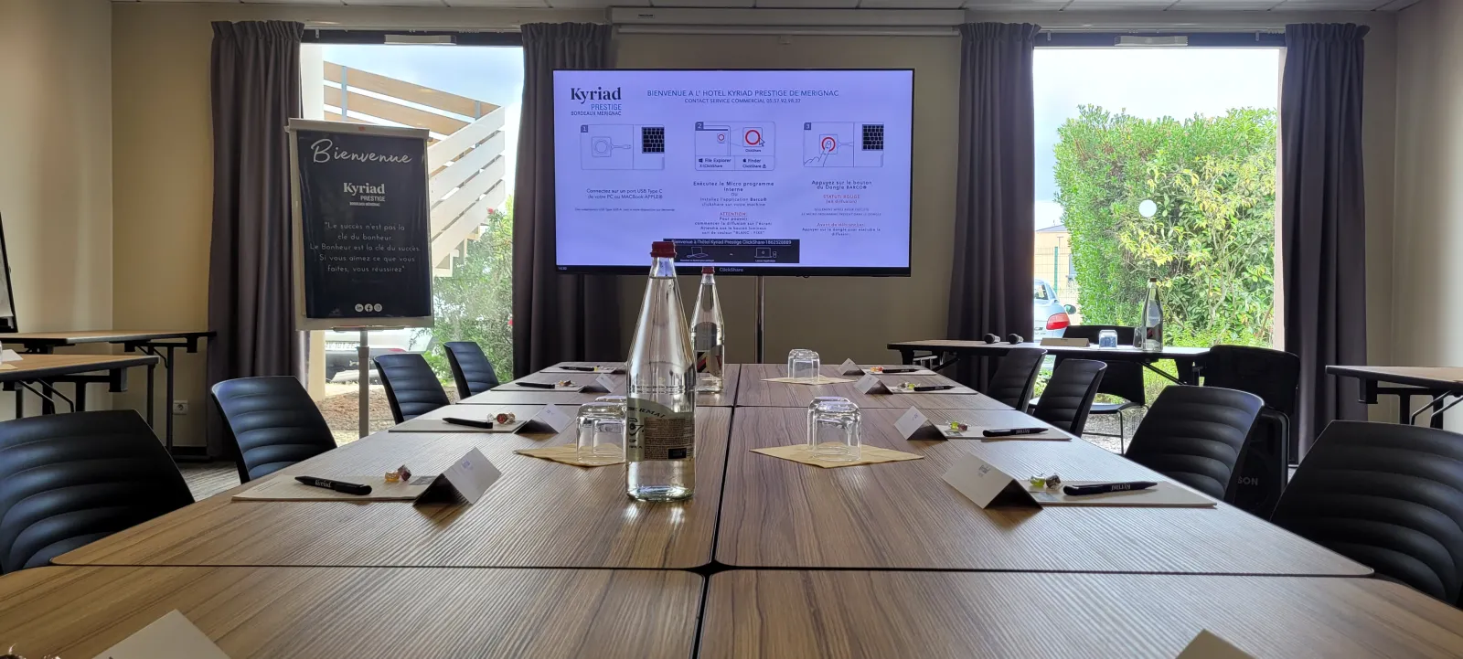 Modern meeting room at Kyriad Prestige Bordeaux Ouest Mérignac featuring a wooden conference table, black chairs, presentation screen, large windows and outdoor greenery views.