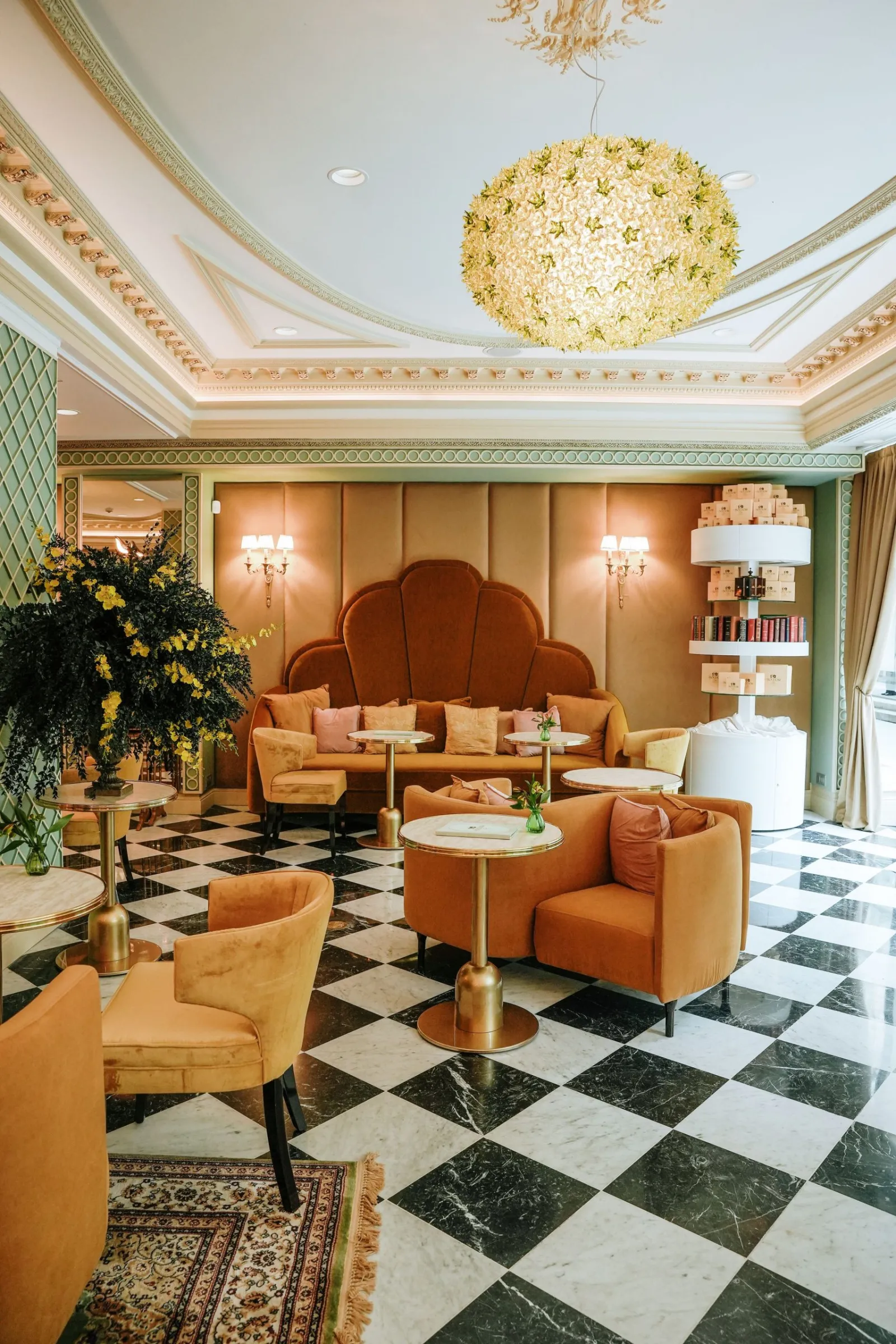 Elegant Art Deco lounge featuring ochre orange furniture, black and white checkered flooring, spherical golden chandelier, sage green and beige walls, yellow floral arrangement and green plants.