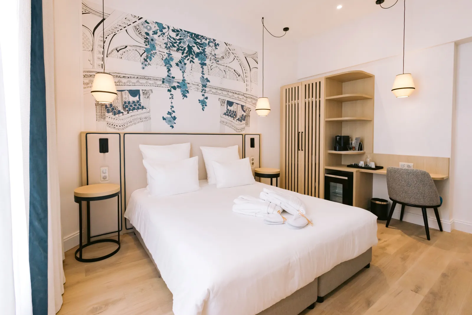 Modern hotel room at Majestic Bordeaux featuring a white bed, natural wood furniture, beige walls, blue botanical artwork above headboard, desk area, open shelving, and wooden flooring.