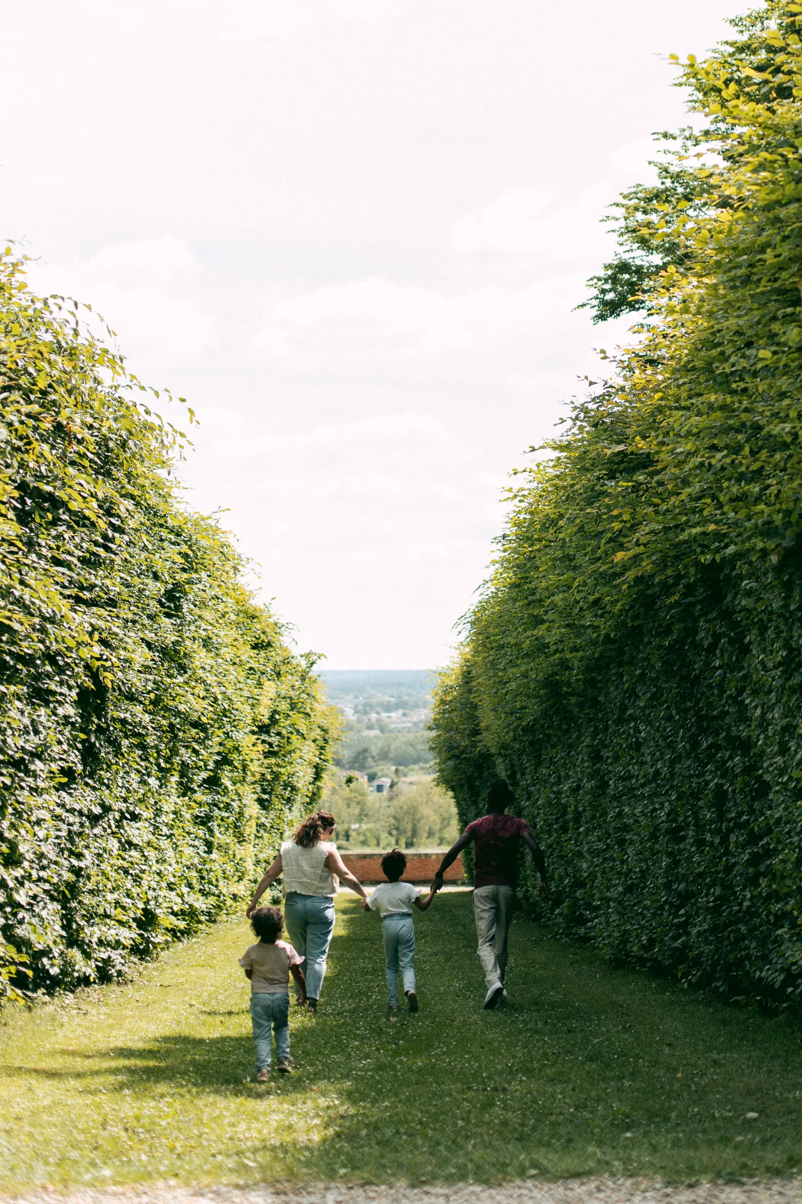 A family of four holding hands and running through a tree-lined pathway with manicured hedges at Malagar Estate, with rolling hills visible in the distance.