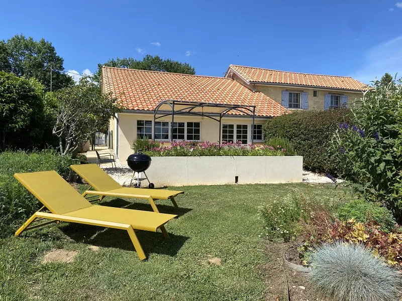 Sunny garden view of Château La Gontrie featuring a yellow lounger, manicured lawn, paved terrace with BBQ, and tile-roofed house in the background.
