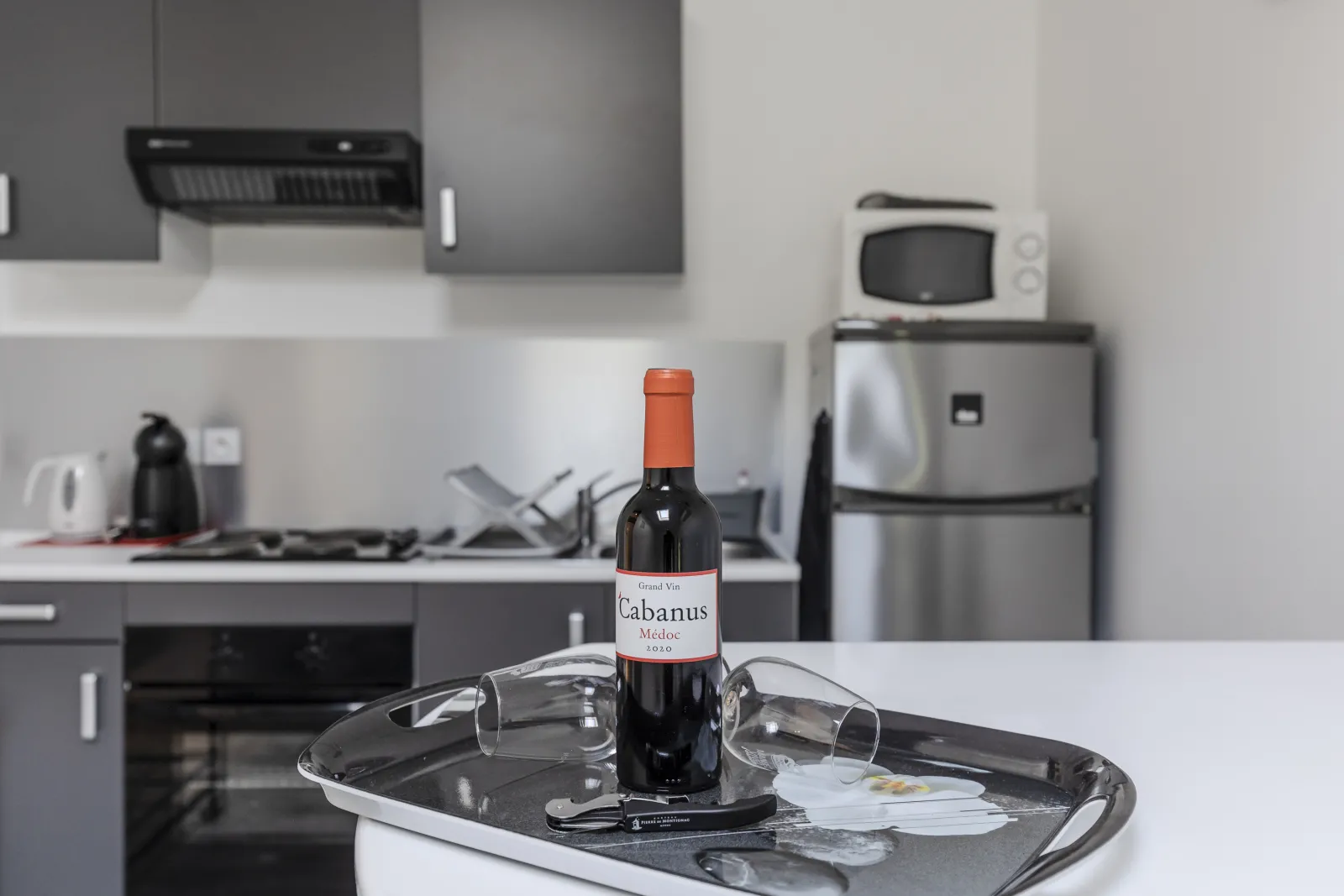 Bottle of red wine Cabanus Médoc 2020 displayed on a black tray with two empty glasses in a modern kitchen with stainless steel appliances.