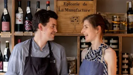 Two smiling staff members in a Bordeaux gourmet shop, standing in front of shelves displaying wines and local products, with the 'Épicerie de La Manufacture' sign in the background.