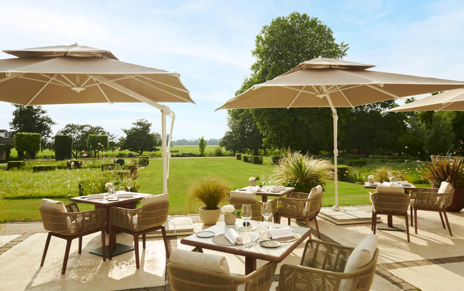 Elegant terrace at Château Hôtel Grand Barrail in Bordeaux featuring set dining tables, wicker chairs, beige umbrellas, and panoramic views of manicured gardens and Bordeaux countryside.