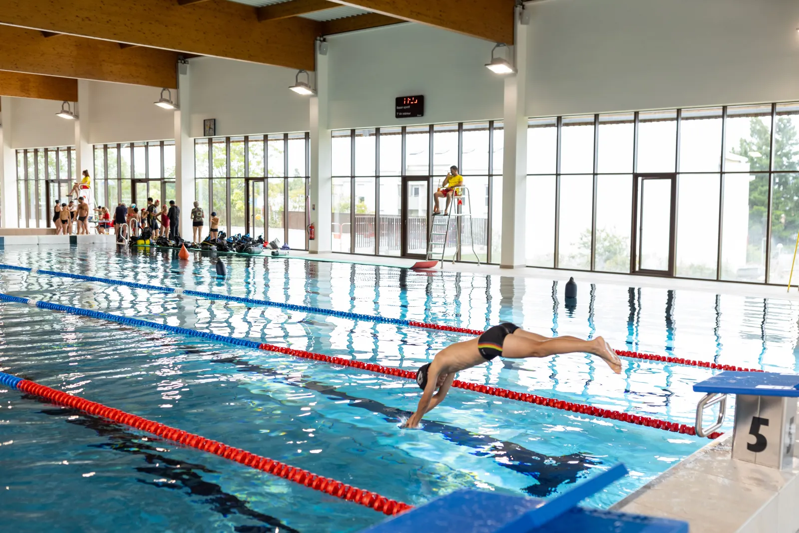 A swimmer performing freestyle stroke in an Olympic pool at the Elodie Lorandi aquatic complex in Bordeaux, with lanes separated by red and blue lane dividers, while a class watches from the poolside.