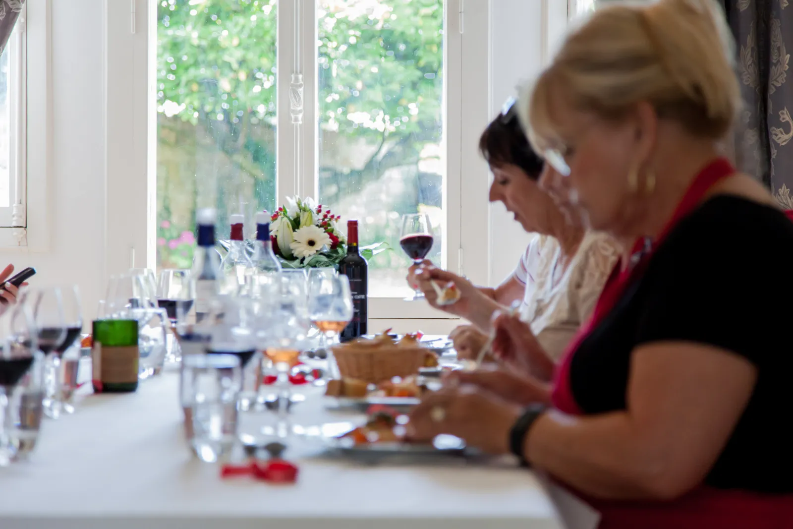 A woman in a black dress with red scarf enjoys red wine during an elegant meal in a bright room with garden views. A table is set with glasses, wine bottles, and a bouquet of white and red flowers.