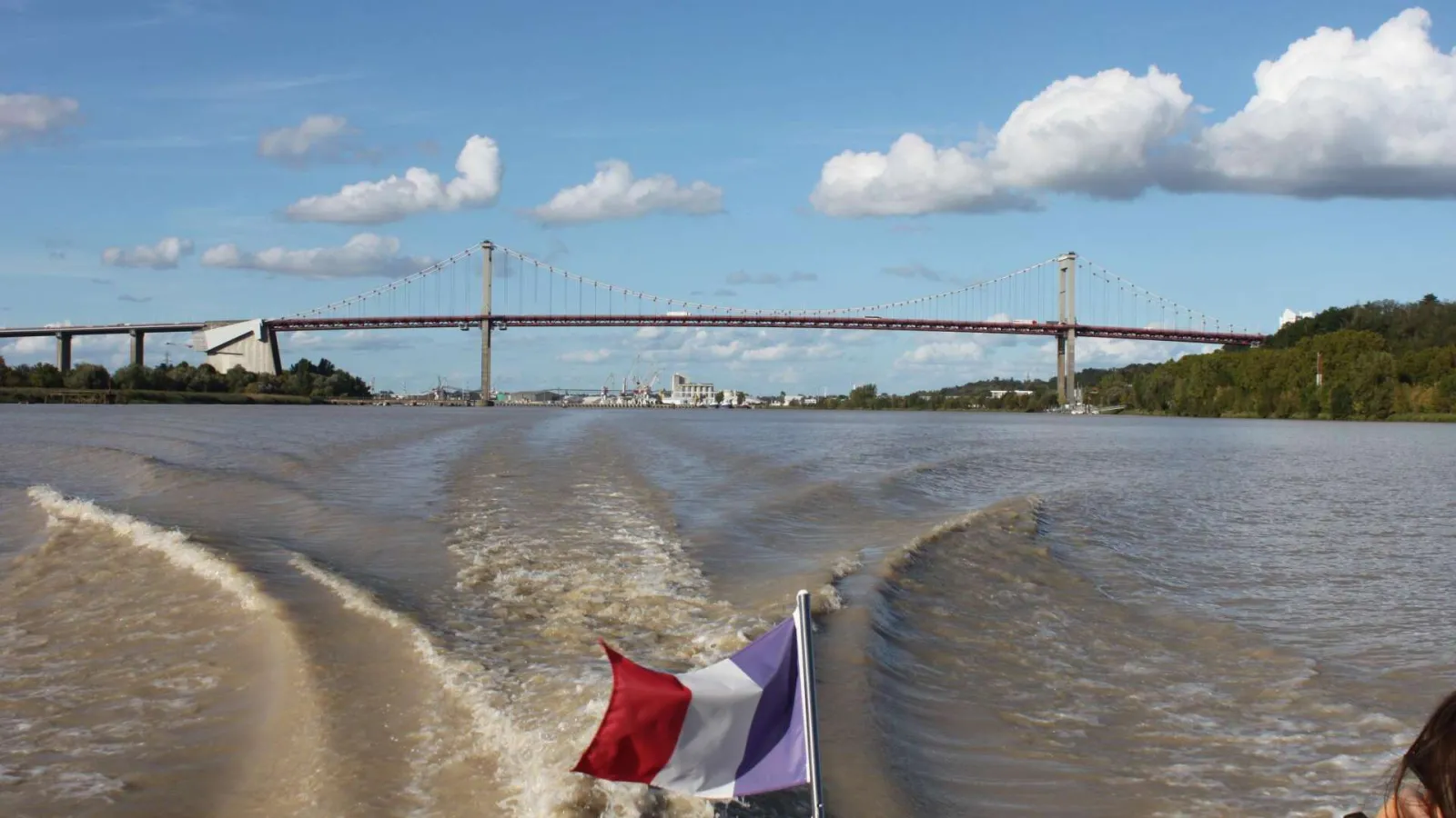 The Aquitaine Bridge spanning the Garonne River in Bordeaux, with the French flag in the foreground and the city skyline in the background under a cloudy blue sky.