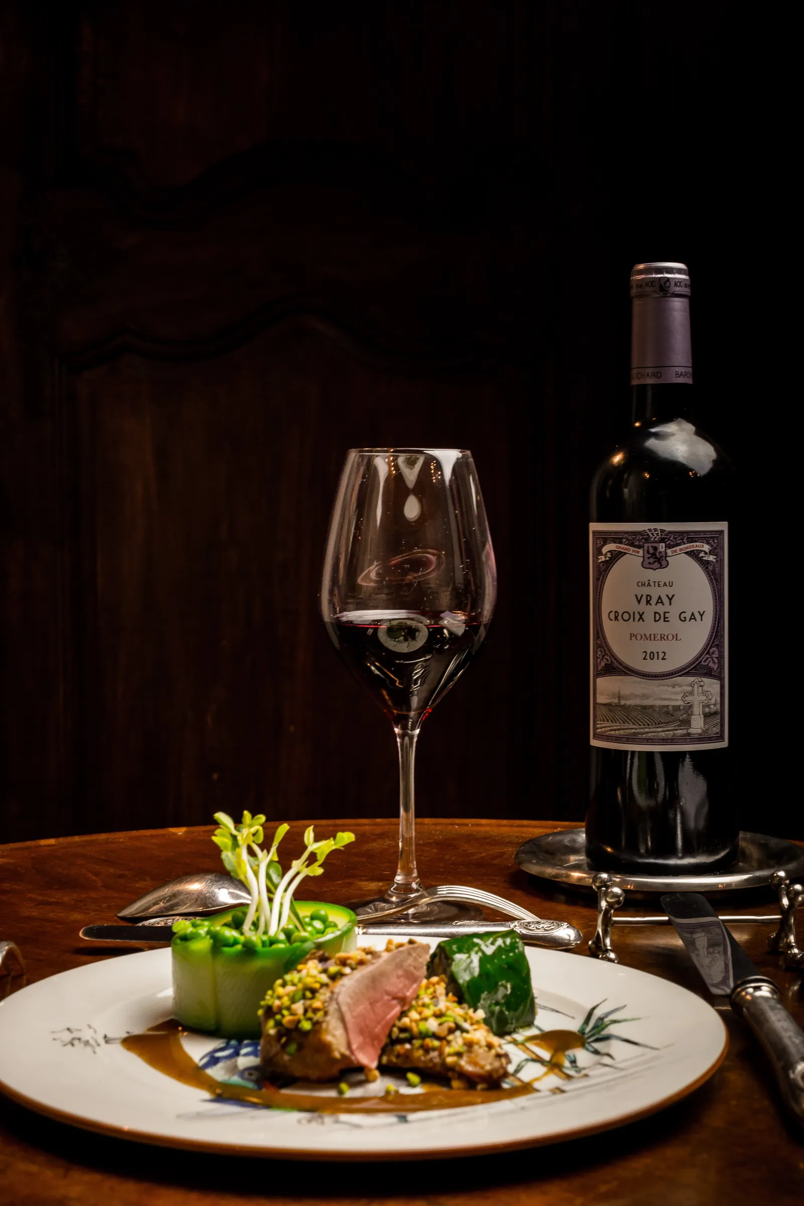 Gourmet plated dish with meat, vegetables and sauce, accompanied by an empty red wine glass and a bottle of Bordeaux Vray Croix de Gay Pomerol 2012 on a wooden table.