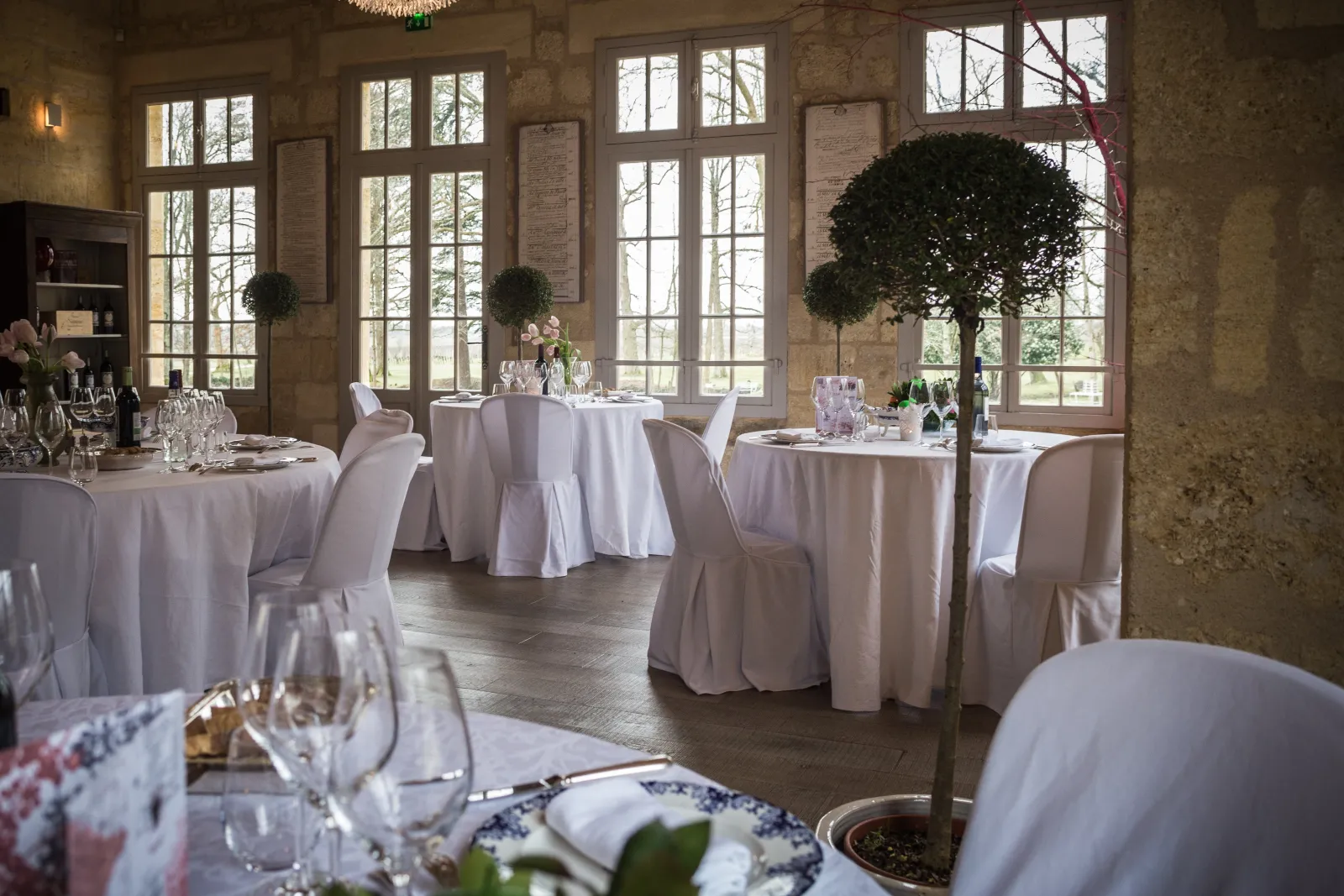 Elegant reception room at La Table de Siaurac in Bordeaux featuring round tables set in white linens, white chair covers, tall arched windows overlooking the grounds, and decorative topiary plants