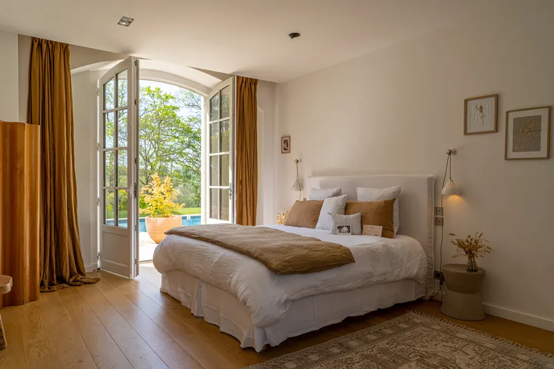 Elegant bedroom at Château Toulouse Lautrec in Bordeaux featuring a white bed, ochre curtains, open French doors overlooking the park and swimming pool