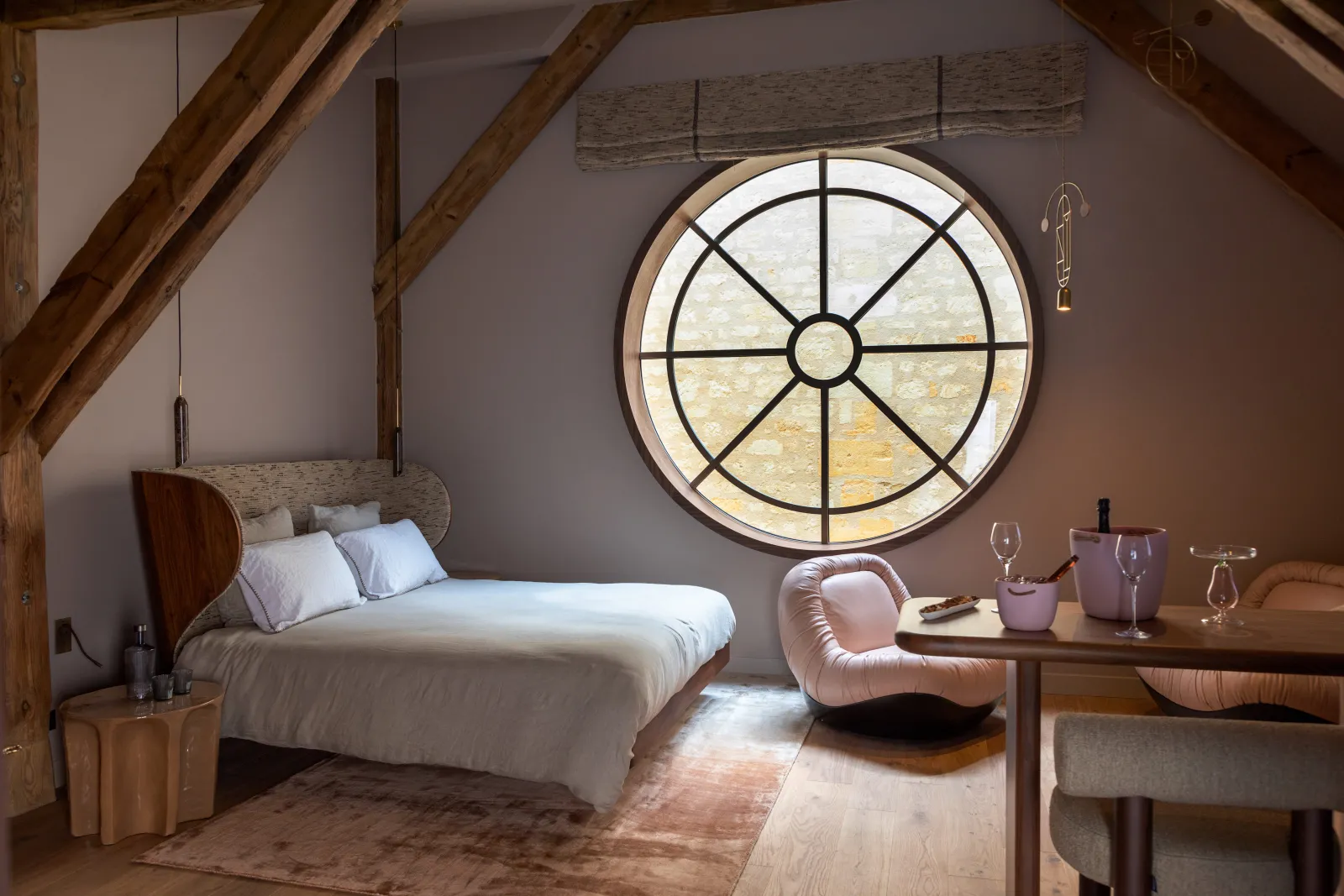 Elegant bedroom at Yndo Hotel in Bordeaux featuring a wooden bed, large circular gridded window, exposed wooden beams, pink armchair, and wooden dining table.