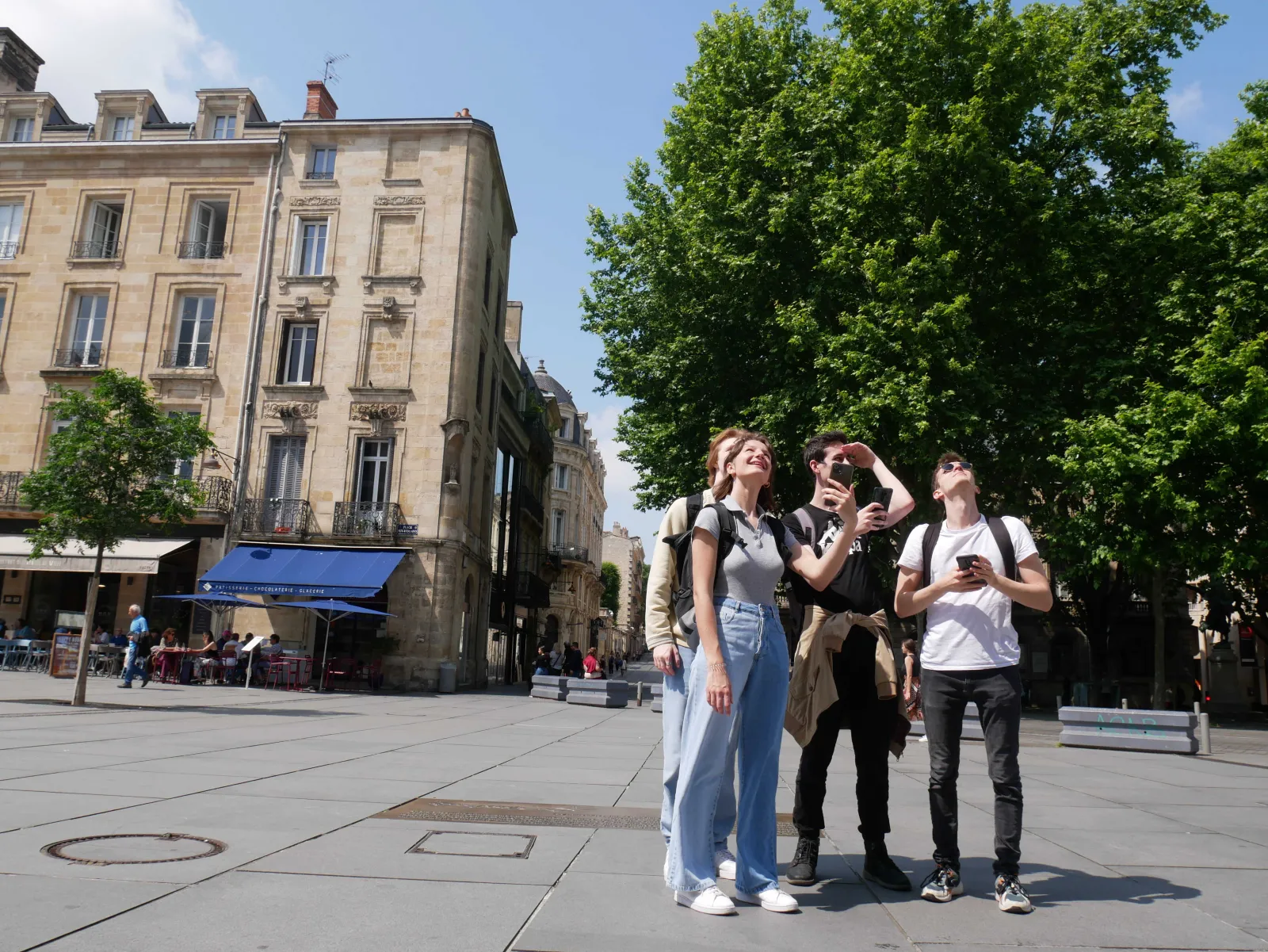 Four young tourists exploring a historic square in Bordeaux, with classical stone buildings and lush green trees, one checking a map.