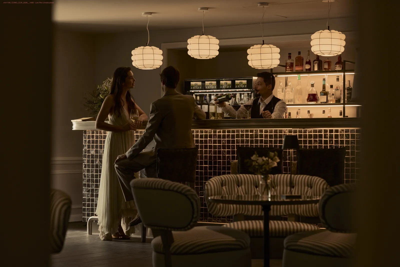 Elegant bar interior with two guests conversing at the counter, a bartender in the background, round pendant lights, striped armchairs in the foreground, and a tiled wall displaying bottles.