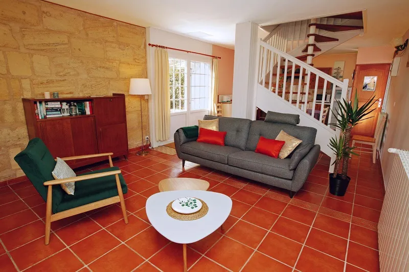 Modern living room at Château La Gontrie in Bordeaux featuring a gray sofa, green armchair, white coffee table, terracotta tiles, yellow stone wall, red cabinet and white staircase