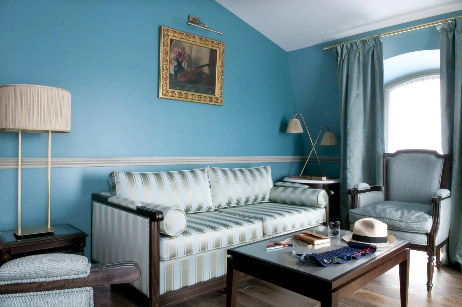 Elegant living room at Hôtel de Sèze in Bordeaux featuring turquoise blue walls, striped white and grey sofa, matching armchair, dark wood coffee table, floor lamp, gilded painting, and grey-green curtains.