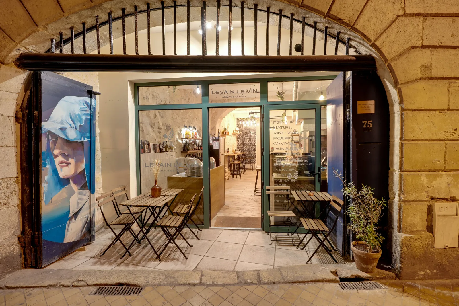 Storefront of Levain Le Vin wine shop in Bordeaux, featuring an outdoor terrace with folding chairs, illuminated display windows and blue promotional poster