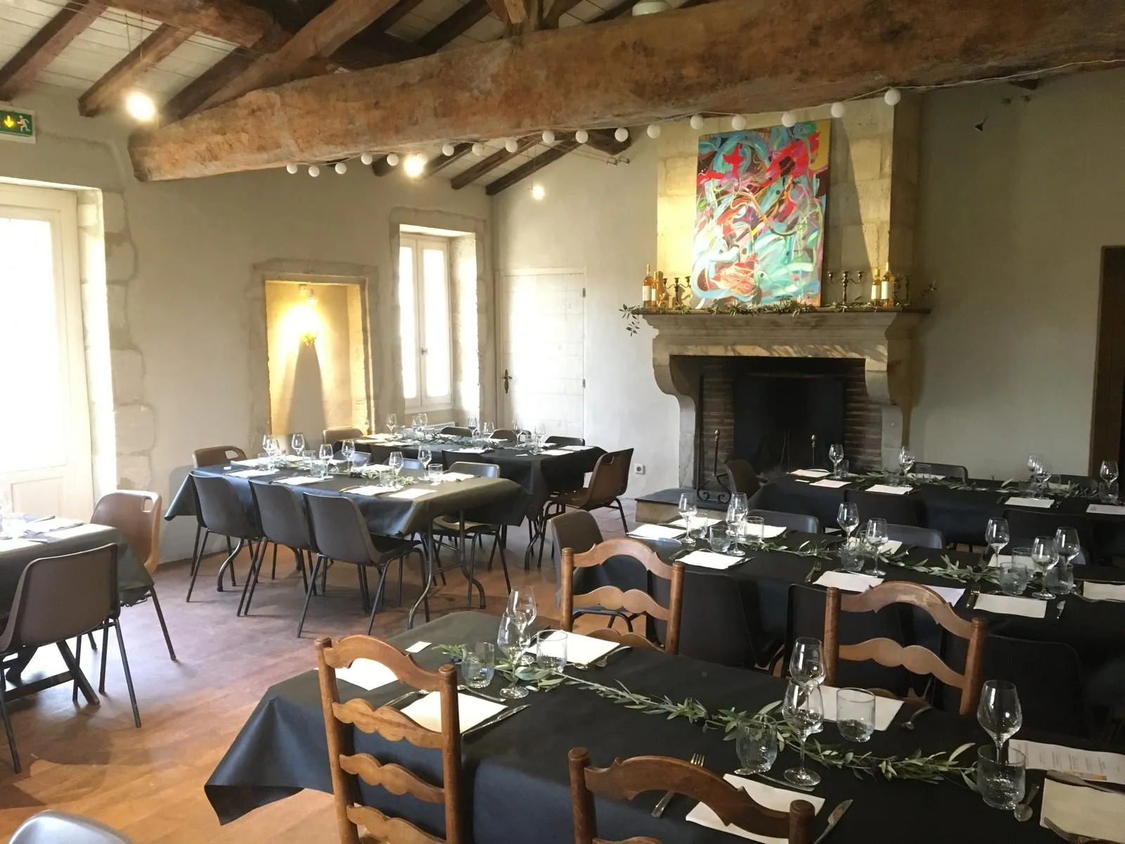 Elegant reception hall at Château Haut-Bergeron in Bordeaux, featuring set dining tables, wooden chairs, historic fireplace, exposed beams and colorful artistic décor