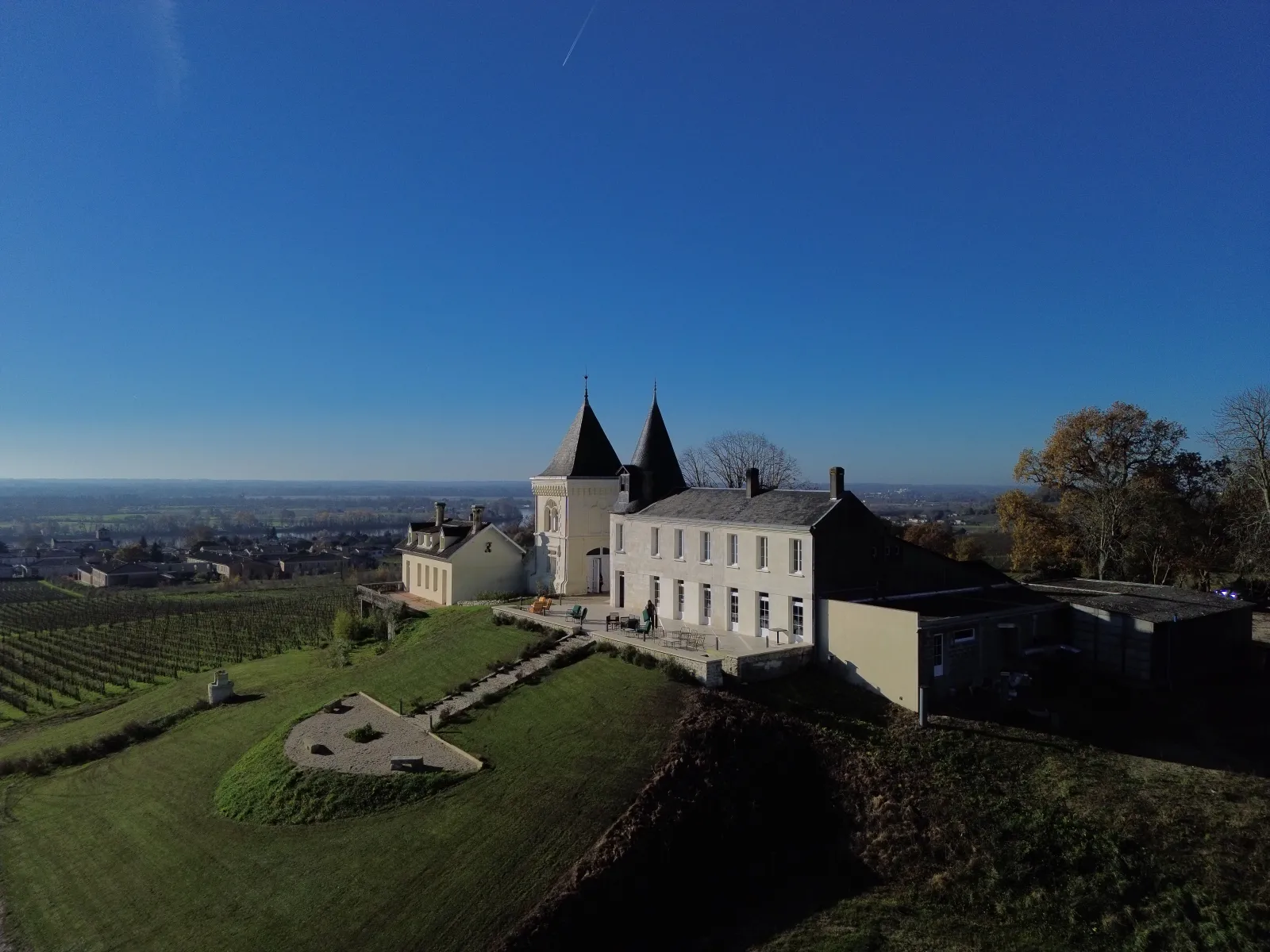 Aerial view of Château CarlMagnus (Maison Roux Oulié) near Bordeaux, a white building with two black pointed towers, surrounded by vineyards and green landscape overlooking the Bordeaux region.