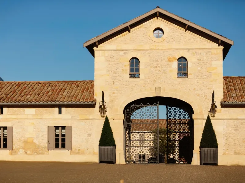 Front facade of Château du Lort in Bordeaux, a stone building featuring a large arched entrance gate, two upper windows, and a peaked roof against a clear blue sky.
