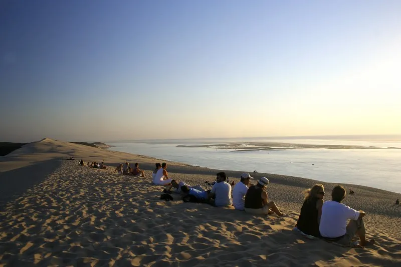Group of visitors sitting on the sand of Dune du Pilat at sunset, overlooking the Arcachon Basin in the background