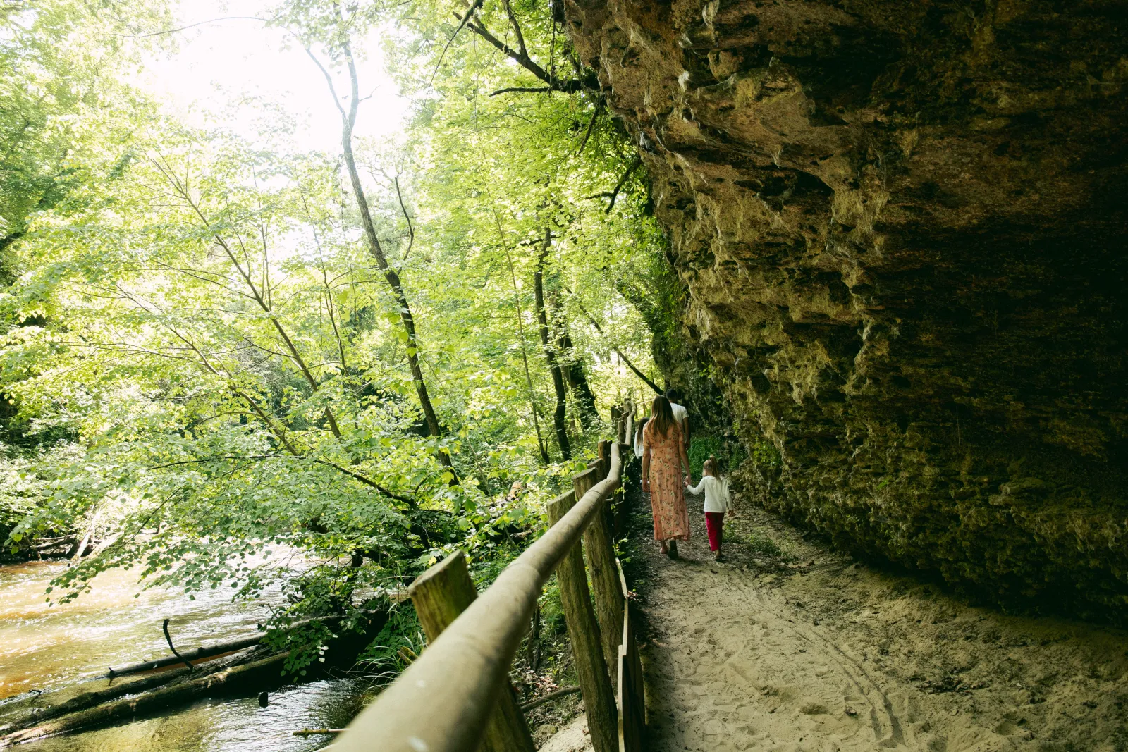 Two people walk along a dirt path beside a river, bordered by a wooden railing and a rocky cliff, surrounded by dense vegetation and trees at the Royal Castle of Cazeneuve.