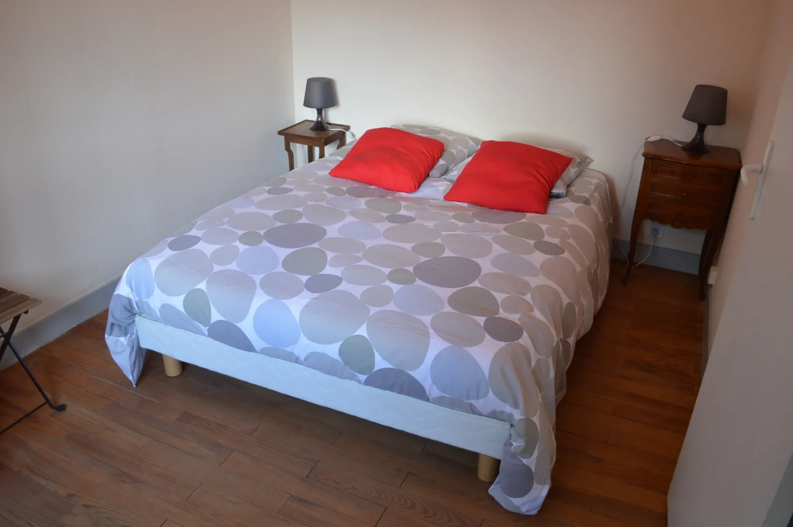 Bedroom with double bed covered with a grey and white patterned duvet, red pillows, wooden bedside tables with lamps, wooden flooring