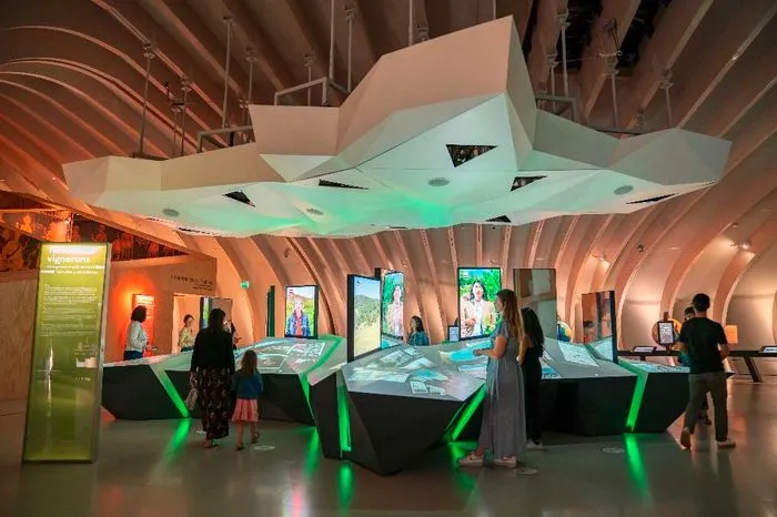 Interactive exhibition at Bordeaux's City of Wine featuring glowing touchscreen displays, an undulating organic-shaped ceiling, and visitors exploring digital installations illuminated in green and orange tones.