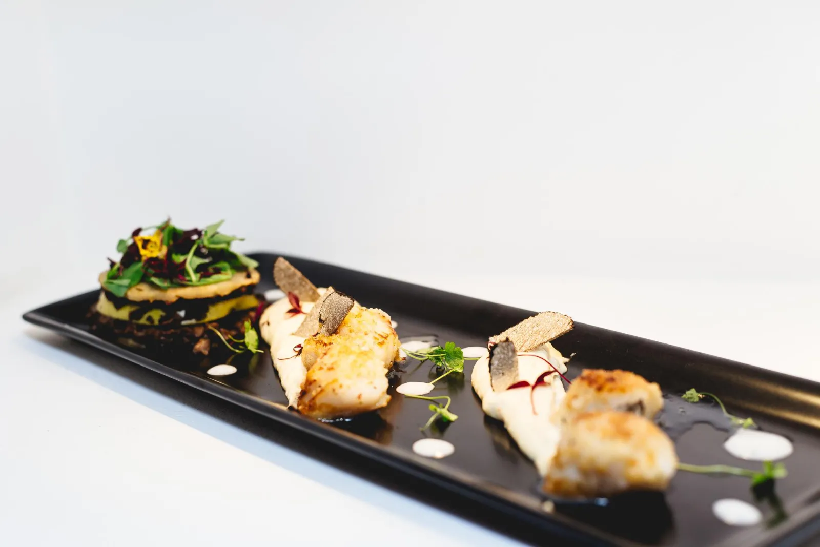 Fine dining plate featuring seared seafood portions topped with truffles, microgreens and sauce arranged on an elongated black slate