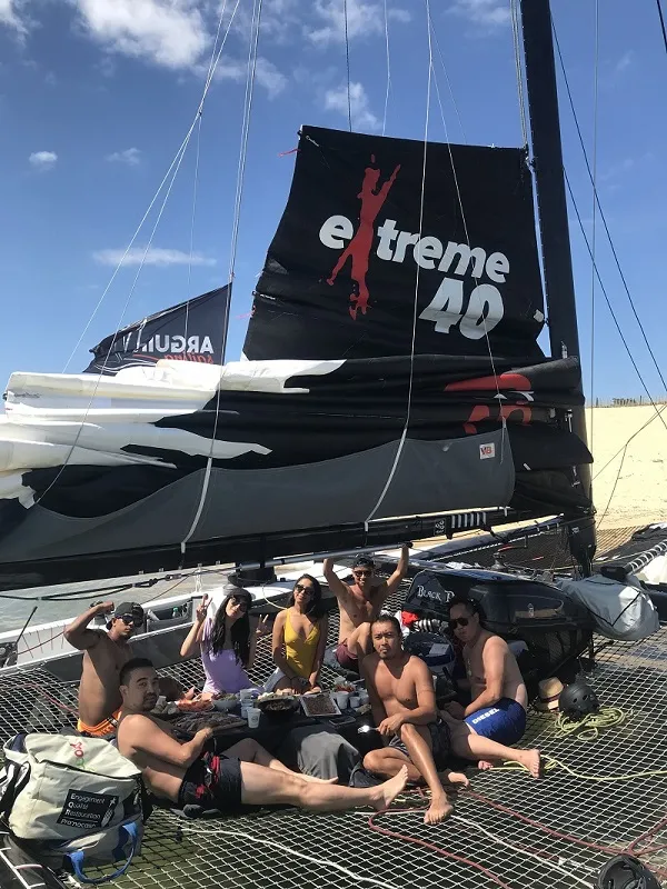 Group of people sitting on the trampoline of a black Extrême 40 catamaran moored in Bordeaux on a sunny day.