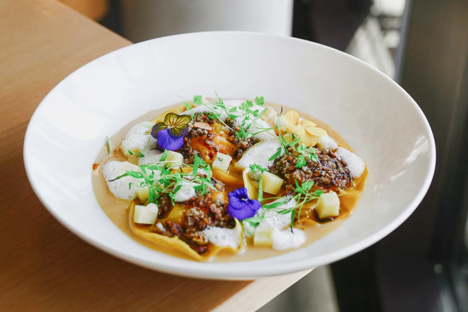 Gourmet plate featuring fresh pasta topped with braised meat, potato cubes, green microgreens, and purple and yellow edible flowers, served in a Bordeaux panoramic restaurant