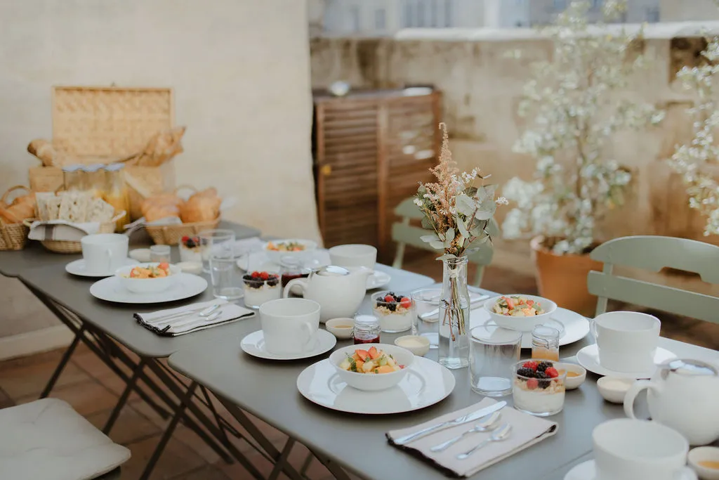 Elegantly set breakfast table with white plates, bowls of fresh fruit, teapots, water glasses and dried flower bouquets in a bright setting in Bordeaux.