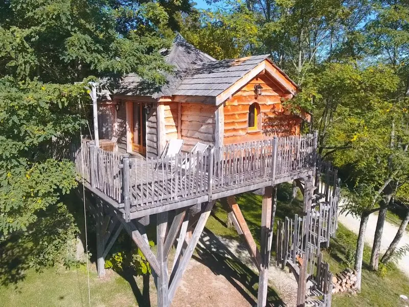 Wooden treehouse on stilts surrounded by green trees, featuring a deck and access stairway, at EcÔtelia Estate in Bordeaux