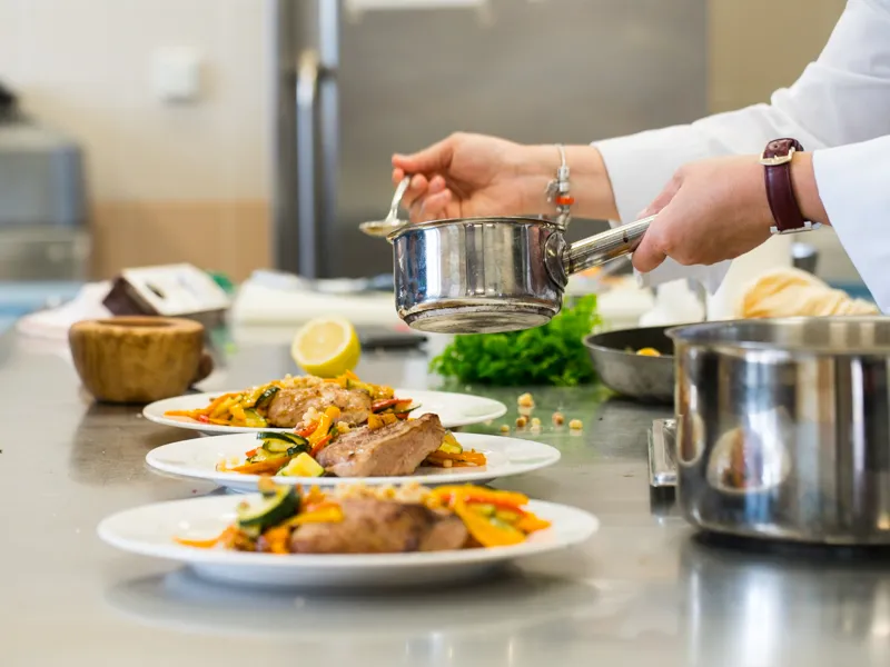 A chef in a kitchen pours sauce from a saucepan over plates of grilled meat and vegetables during a cooking workshop at Château Kirwan in Bordeaux.